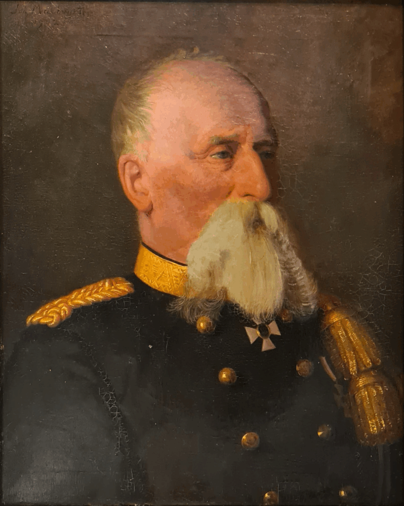 Portret in driekwart van een man met grote witte snor en baard, in zwart uniform met dubbele rij gouden knopen.