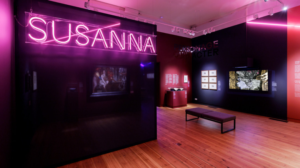 In een ruimte staat hoog tegen een zwarte wand in grote roze neonletters het woord Susanna. Achterin de ruimte zijn enkele kunstewerken te zien met het thema Susanna. In het midden van de ruimte staat een bankje