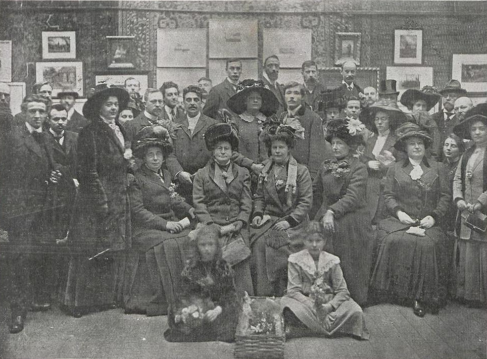 Op een zwart-wit foto poseert een grote groep mensen in een zaal gevuld met schilderijen. Vooraan zitten twee jonge meisjes op de grond. Daarachter zitten vijf vrouwen met hoeden en gekleed in dikke jassen. Naast en achter hen staan nog eens ongeveer 25 mensen, waaronder nog eens 8 vrouwen, de rest mannen.